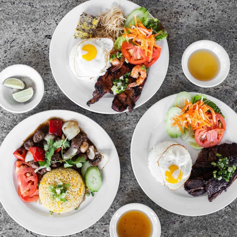 Three Vietnamese dishes with grilled meats, fried eggs, rice, and fresh vegetables on a dark table.